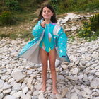 A girl on a pebble beach giving a thumbs up, wearing a UV Protective Long Sleeve Swimsuit Green Ice Cream and matching robe.